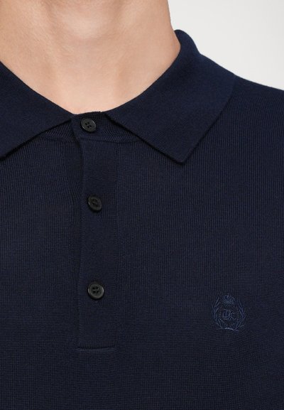 Polo din navy, cu textură canelată, cu guler clasic și patru butoane negre. Include un logo brodat subtil în aceeași culoare.
