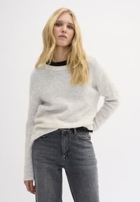 Maglione grigio chiaro con una texture, scollo rotondo, indossato sopra una maglietta nera, abbinato a jeans scuri a vita alta.