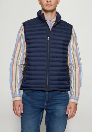 Mand iført mørkeblå quiltet vest med lynlås over en langærmet, stribet skjorte og blå jeans, stående foran en ensfarvet lys baggrund.