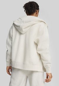 Felpa bianca in pile con cappuccio e polsini a coste, caratterizzata da una consistenza morbida e una vestibilità over size. La vista posteriore mostra una silhouette rilassata.