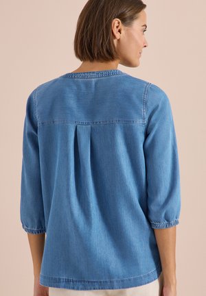 Femme aux cheveux bruns courts portant une chemise en denim bleu clair avec des manches trois-quarts, de dos sur un fond beige.