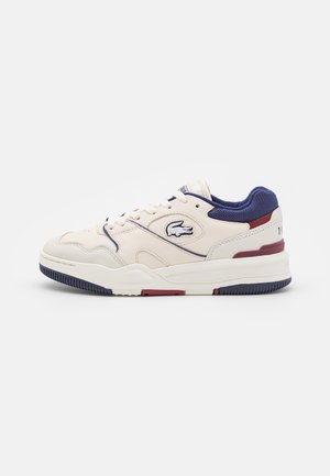 Niski biały sneaker z granatowymi i czerwonymi akcentami, teksturowaną podeszwą, sznurowaniem z przodu i logo krokodyla po boku.