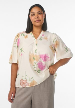 Woman with straight black hair wearing a cream blouse with pastel floral print and beige pants, standing with one hand on hip.