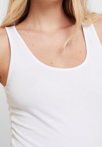 Débardeur blanc en coton avec un col rond et des fines bretelles. Le tissu est lisse et ajusté, révélant subtilement le teint de la peau en dessous.