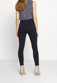 Mujer vestida con jeans ajustados oscuros, una blusa sin mangas de rayas verticales en blanco y negro, y sandalias plateadas de tacón alto, vista desde atrás.