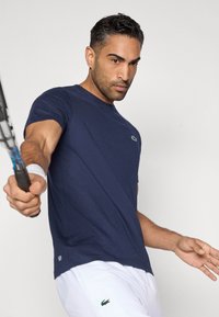 Granatowy t-shirt z okrągłym dekoltem, wykonany z lekkiego materiału, z zielonym logo na piersi, w połączeniu z białymi szortami. Trzyma rakietę tenisową.