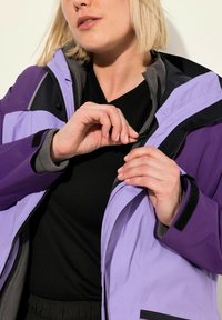 Veste imperméable mauve et lavande avec un zip à l'avant, doublure grise et accents noirs, portée sur un haut noir. Détail de zip horizontal visible.