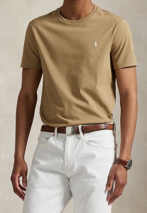 Man draagt een beige T-shirt met korte mouwen met klein wit logo, witte broek, bruine leren riem en bijpassend horloge op een neutrale achtergrond.