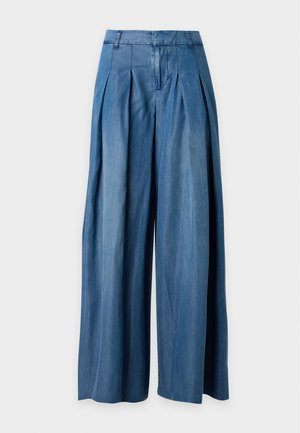 Femme debout les mains dans les poches, portant un t-shirt à manches courtes rayé bleu marine et blanc, un pantalon bleu à jambes larges et des chaussures blanches.