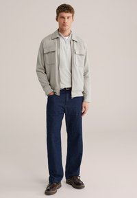 Veste zippée gris clair avec deux poches poitrine, associée à une chemise blanche unie et un jean droit bleu foncé. Chaussures à lacets marron.