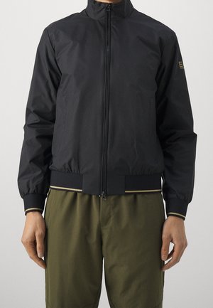 Veste bomber noire à fermeture éclair avec poignets et ourlet côtelés présentant de fines rayures dorées, portée avec un pantalon vert olive.