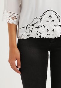 Blouse blanche avec ourlet en dentelle complexe, présentant des bords festonnés et des motifs floraux. Le tissu est lisse ; porté sur un jean noir ajusté.