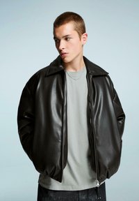 Giacca bomber in ecopelle nera con un collo ampio, chiusura con zip e maniche larghe. Indossata sopra una t-shirt grigia chiara, su uno sfondo neutro.