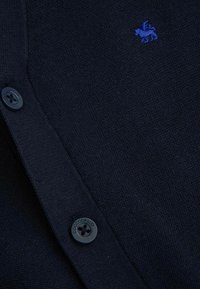 Granatowy kardigan wykonany z teksturowanej dzianiny o gładkim wykończeniu. Posiada ciemny guzik i małe niebieskie logo lwa.