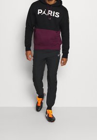Sweat à capuche noir et bordeaux avec le texte « PARIS » et le logo Jumpman, associé à un pantalon noir Nike et des crampons noirs avec des accents orange.