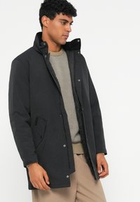 Μαύρο παλτό με φερμουάρ και κούμπωμα, με ψηλό γιακά και πλαϊνές τσέπες, τοποθετημένο πάνω από ένα ανοιχτό καφέ υφασμάτινο πουλόβερ.