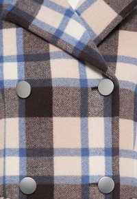Manteau en laine à carreaux affichant un motif beige, marron et bleu avec des boutons argentés sur un design croisé. Texture douce.