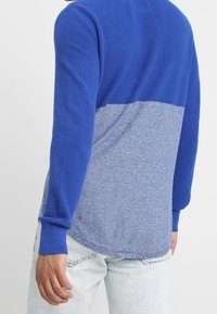 Chemise en tricot à manches longues avec une section supérieure bleue vive, un tissu texturé et une section inférieure bleu clair chiné avec un ourlet incurvé.