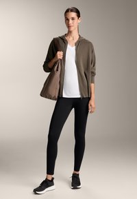 Olivově zelená mikina na zip, bílý tričko s výstřihem do V, černé legíny a černé tenisky. Hnědá kabelka přes rameno s hladkou texturou.