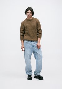 Joven de pie con un suéter marrón texturizado, jeans sueltos de color azul claro y zapatos negros de suela gruesa, contra un fondo blanco liso.