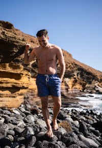 Blauwe zwemboxers met sterrenpatroon, tailleband met trekkoord en zijzakken; model staat op een rotsachtig strand zonder shirt, blootsvoets, glimlachend.