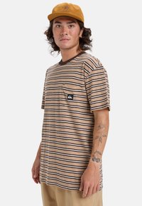 Camiseta de manga corta a rayas en color beige con líneas marrones, con un bolsillo en el pecho y un parche de logo. Se lleva con pantalones de color claro y una gorra.