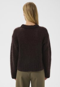 Tumši brūns adīts džemperis ar platu ribotu apkakli un vaļēju piegriezumu. Materiāls izskatās mīksts un pūkains, ar teksturētu virsmu.