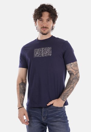 Hombre con cabello rizado que lleva una camiseta azul marino con un patrón abstracto blanco, de pie con una mano en el bolsillo y tatuajes en ambos brazos.