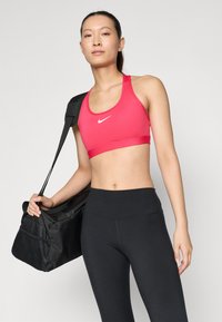 Sostén deportivo rosa con el logo de Nike, combinado con leggings negros de tiro alto. La modelo lleva un bolso negro con un bolsillo de malla.