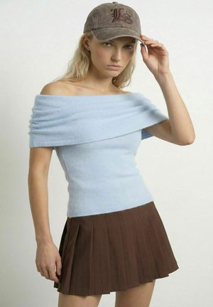 Ung kvinde iført en lyseblå off-shoulder top, brun plisseret nederdel og grå kasket, der rører ved kasketkanten med et neutralt udtryk.