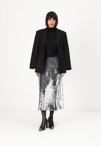 Zwarte oversized blazer over een doorschijnende zwarte top, gecombineerd met een zilveren rok van pailletten tot de knie. Zwarte gepatineerde hakken en doorschijnende panty's maken de outfit compleet.