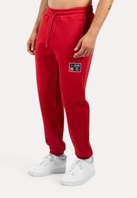 Pantalones de jogging de felpa roja con cintura elástica, bolsillos laterales y un parche de logo prominente que incluye texto. Combinados con zapatillas blancas.