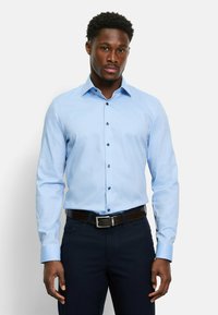 Hellblaues Langarmhemd mit Knopfleiste, ausgestattet mit dunklen Knöpfen, einem spitzen Kragen und einer glatten Stofftextur. Kombiniert mit dunkler Hose.