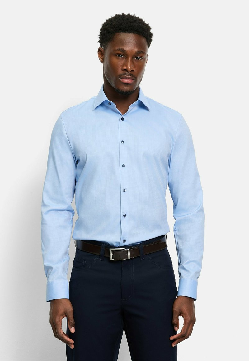 Hellblaues Langarmhemd mit Knopfleiste, ausgestattet mit dunklen Knöpfen, einem spitzen Kragen und einer glatten Stofftextur. Kombiniert mit dunkler Hose.