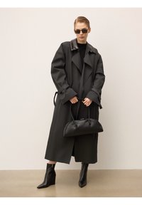 Nainen, jolla on ylleen suuri tumma trench-coat, mustat saappaat ja suorakulmaiset aurinkolasit, piti mustaa nahkabagia beigeä taustaa vasten.