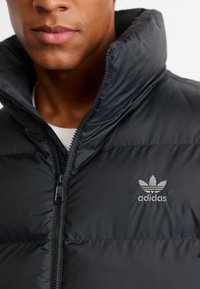 Sort puffet jakke med høj krave, lynlås og reflekterende Adidas-logo. Har quiltet tekstur og glat stof.