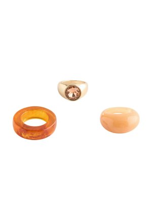 Three rings on white background: one gold with large brown gemstone, one translucent amber, and one smooth beige resin band.