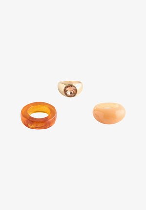 Three rings on white background: one gold with large brown gemstone, one translucent amber, and one smooth beige resin band.