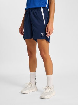 Person trägt dunkelblaue Sportshorts mit weißen Seitenstreifen und weißen Turnschuhen, steht vor einem einfarbigen Hintergrund.