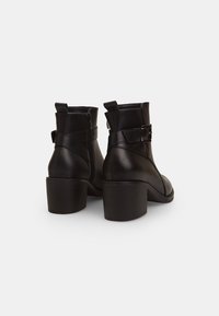 Botas de tobillo de cuero negro con tacón grueso, cremallera lateral y una correa con hebilla en el tobillo para un detalle adicional.