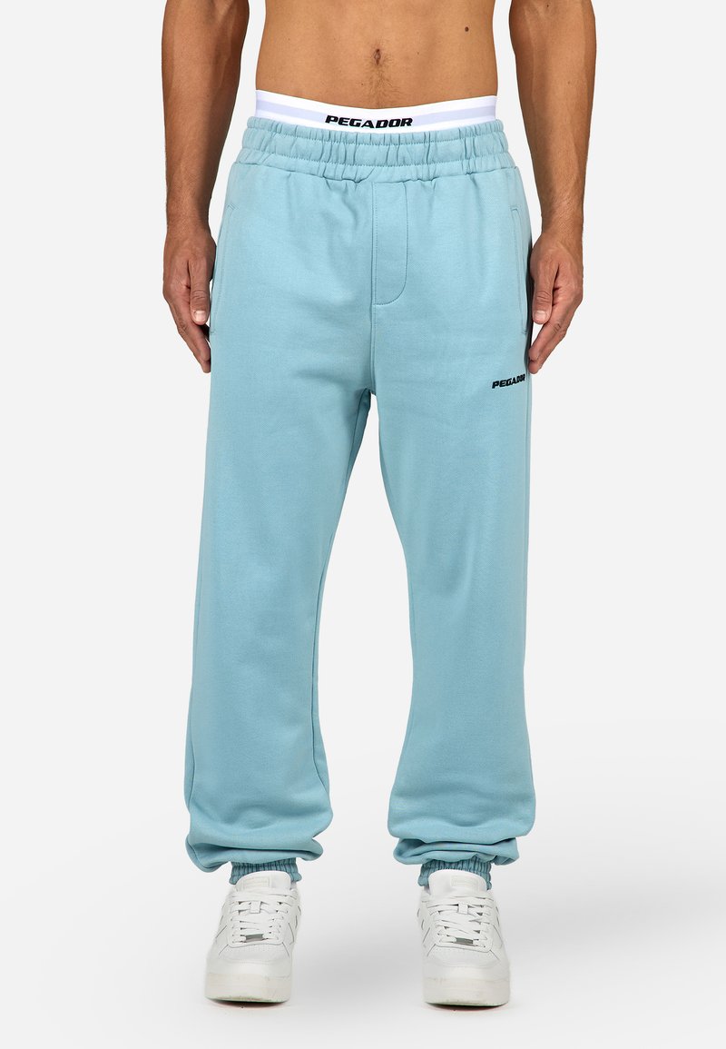 Šviesiai mėlynos sportinės kelnės su elastiniu juosmeniu, turinčios šonines kišenes ir gipiūro tamprias rankoves. Juodas "PEGADOR" logotipas kairiame šlaunyje.
