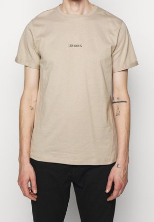 T-shirt en coton beige avec un col rond et des manches courtes. Présente un texte noir "LES DEUX" centré sur la poitrine. Design simple, coupe décontractée.