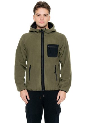 Un homme porte une veste polaire à capuche vert olive, avec une fermeture éclair noire sur le devant et des poches, assortie à un pantalon noir sur fond blanc.