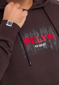 Brązowa bluza z kapturem z czerwono-białym napisem "BKLYN NEW YORK CITY", zdobiona fakturowanym wzorem i sznureczkami do ściągania. Etykieta na rękawie.