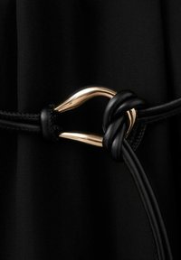 Primo piano di un cordoncino in pelle nera annodato attorno a una fibbia in metallo dorato lucido su sfondo di tessuto nero.