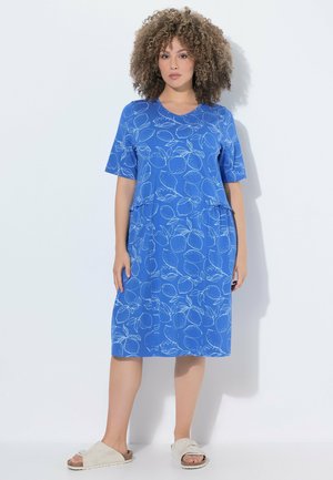 Donna con capelli ricci che indossa un vestito blu fino al ginocchio con motivo di limoni e sandali beige, in piedi su uno sfondo bianco.