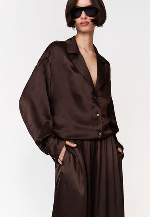 Žena v nadrozmernej tmavohnedej saténovej blúzke a zodpovedajúcich širokých nohaviciach, s čiernymi slnečnými okuliarmi, ruky v vreckách, na bielom pozadí.