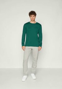 Camicia verde a maniche lunghe in un tessuto morbido, abbinata a pantaloni grigi chiari. La camicia presenta un piccolo logo e un design dalla vestibilità rilassata.