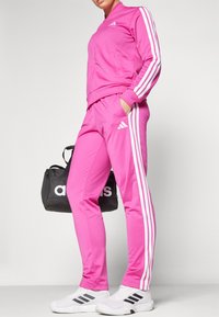 Pink tracksuit with white side stripes, featuring a zip-up jacket and matching pants. Accompanied by a black duffel bag with "adidas" logo.