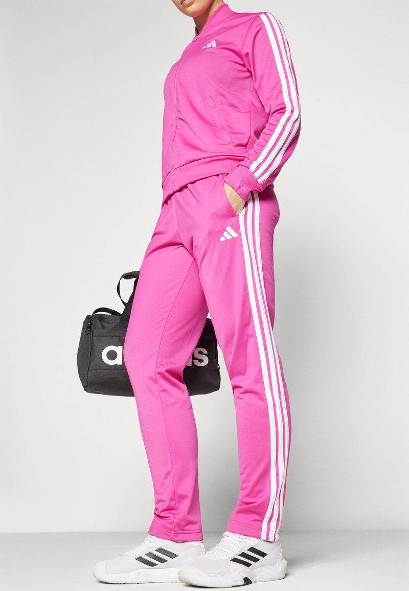 Chándal rosa con rayas blancas a los lados, que incluye una chaqueta con cremallera y pantalones a juego. Acompañado de un bolso de deporte negro con el logo de "adidas".